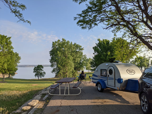 Lakeside campsite featuring a T@G teardrop RV rental, picnic table, and person relaxing by the water.