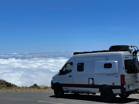White Winnebago Revel 4x4 camper van on a mountain road, high above clouds. Blue sky. Epic rv rental adventure awaits!