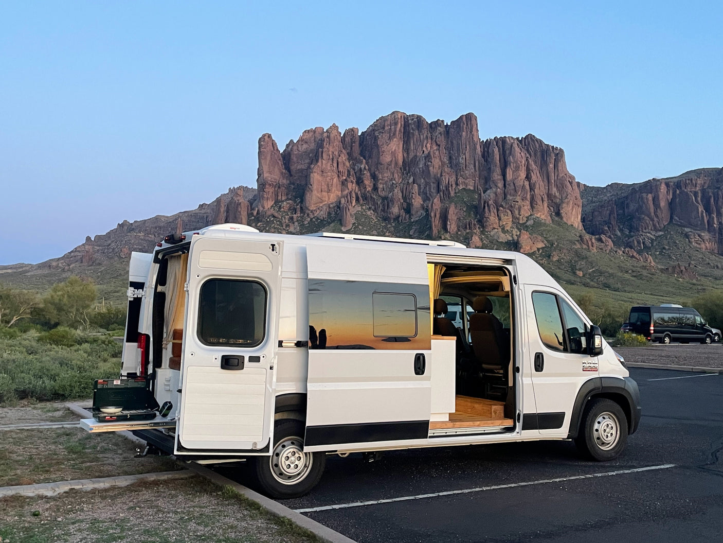 An RV rental, a white campervan with open doors, parked in front of majestic mountains, showcasing its outdoor kitchen.