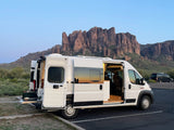 An RV rental, a white campervan with open doors, parked in front of majestic mountains, showcasing its outdoor kitchen.