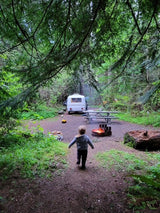 Family camping fun! Child approaches an RV rental campsite with a warm campfire in a lush forest.