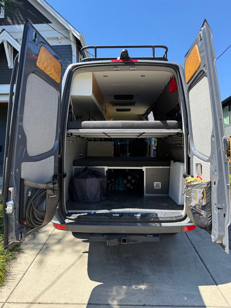 Rear of a gray RV rental camper van with open doors, revealing a cozy bed and organized gear storage for adventures.