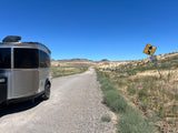 Silver RV rental trailer on a dirt road in a sunny, arid landscape with distant hills and signs.