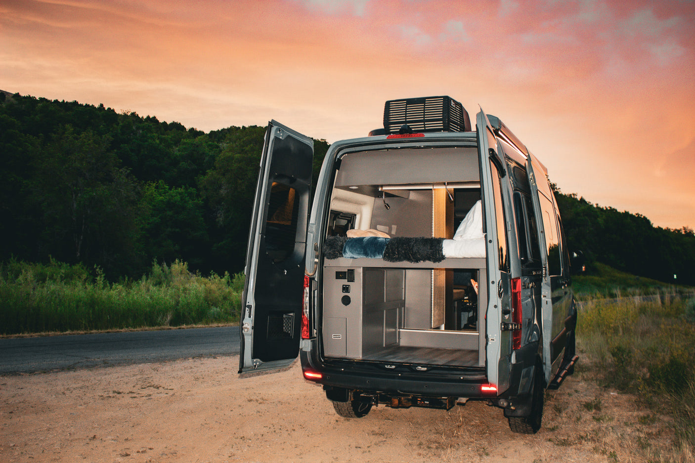 Stylish camper van RV rental with open rear doors showing bed & storage, parked on a dirt road at sunset.