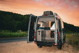 Stylish camper van RV rental with open rear doors showing bed & storage, parked on a dirt road at sunset.