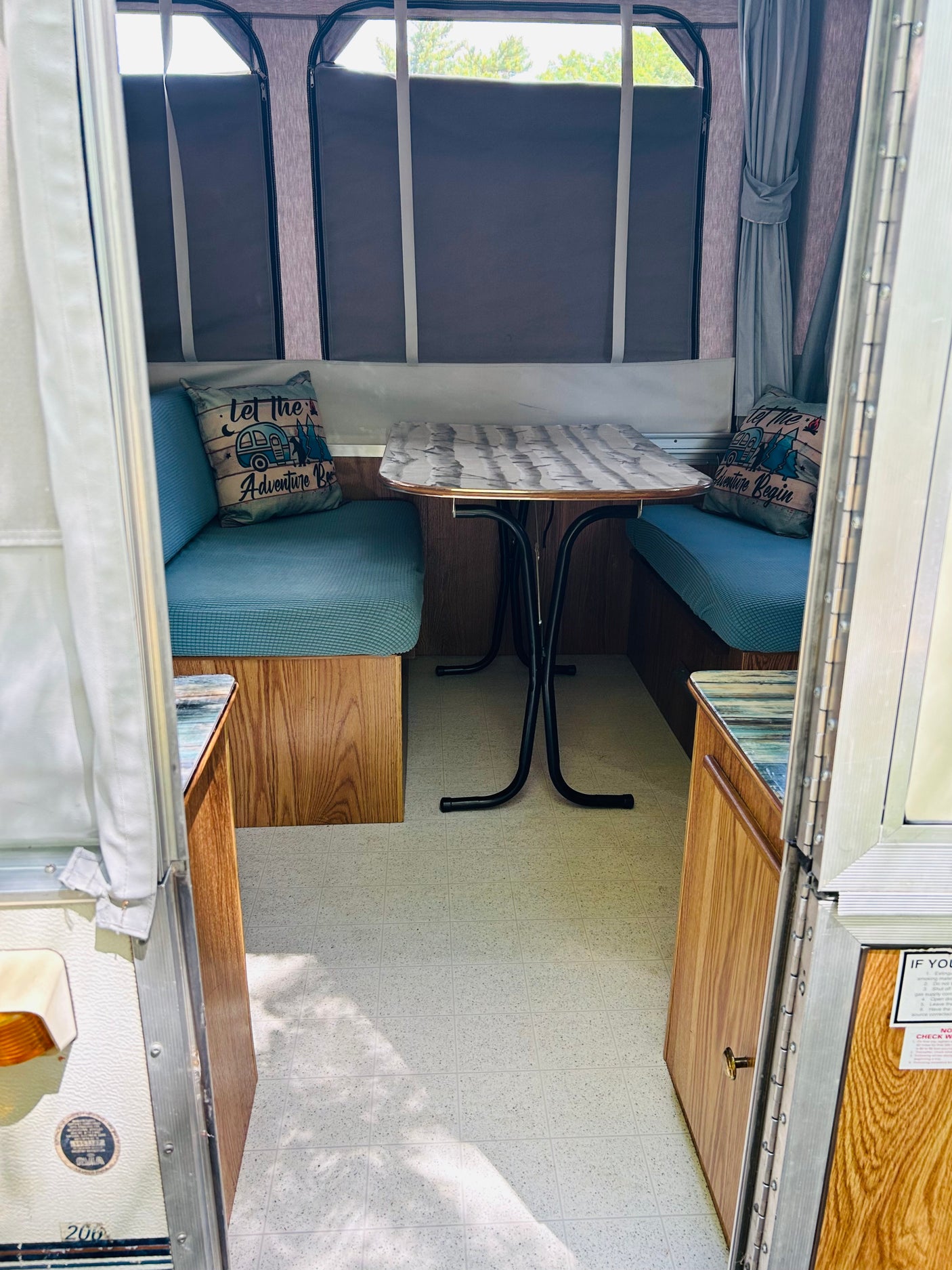 Inside a cozy pop-up rv rental: blue-cushioned dining benches, a table, and "Let the Adventure Begin" pillow.