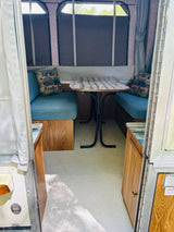 Inside a cozy pop-up rv rental: blue-cushioned dining benches, a table, and "Let the Adventure Begin" pillow.