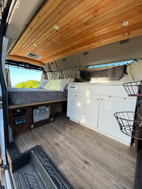 Modern RV rental interior featuring a comfy bed, kitchen, wood ceiling, and scenic window views.