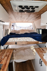 Cozy RV rental: elevated bed with blue blanket, wooden plank ceiling, AC unit, wood counters, and scenic window view.