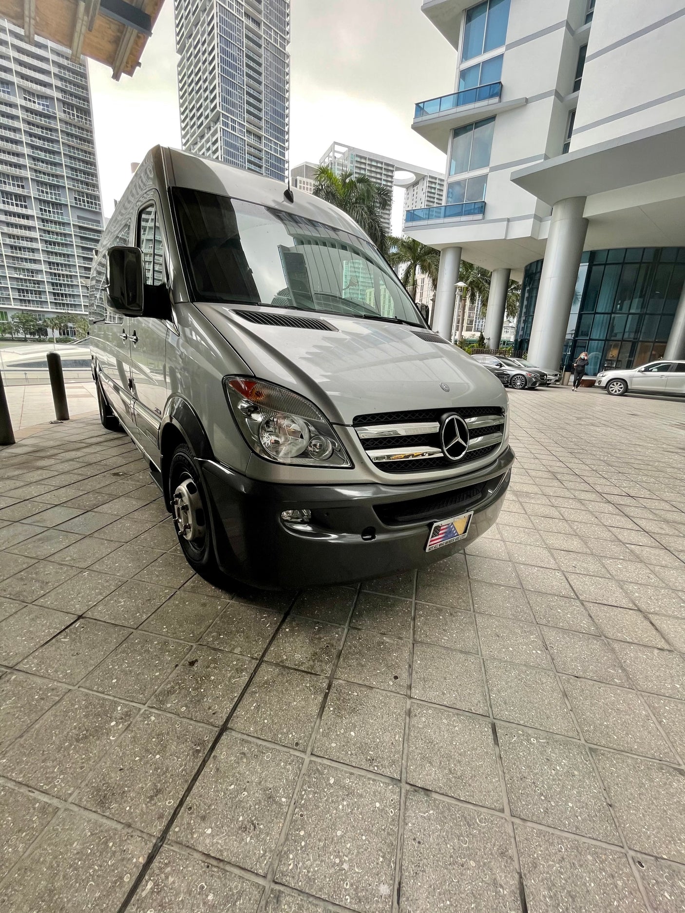 A silver Mercedes-Benz Sprinter van, ideal for RV rental, parked on patterned grey tiles. Tall buildings frame the urban scene.