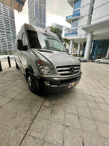 A silver Mercedes-Benz Sprinter van, ideal for RV rental, parked on patterned grey tiles. Tall buildings frame the urban scene.