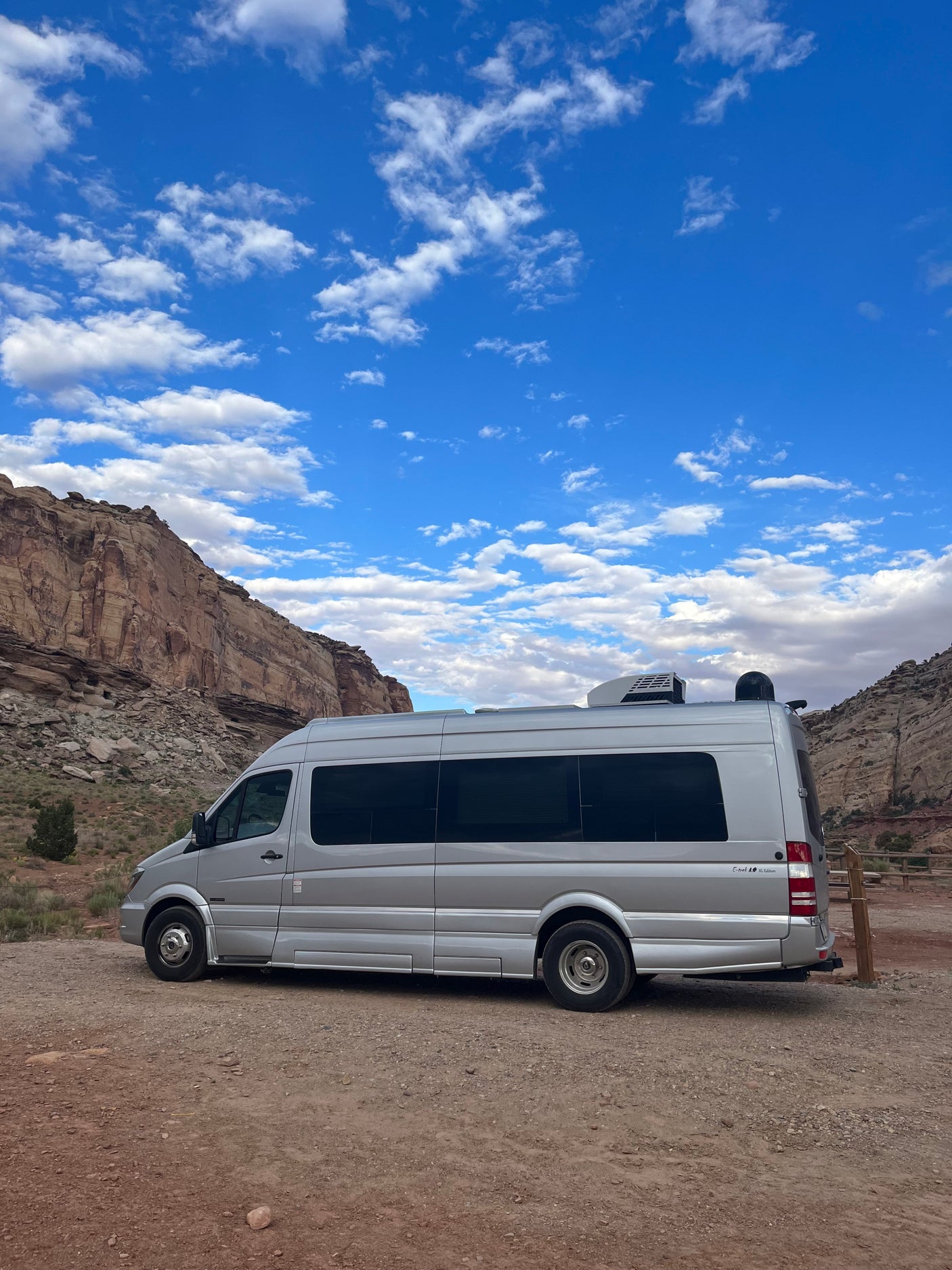A silver camper van, possibly an rv rental, sits on a dirt road, framed by majestic desert mountains under a clear blue sky.