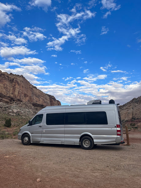 A silver camper van, possibly an rv rental, sits on a dirt road, framed by majestic desert mountains under a clear blue sky.