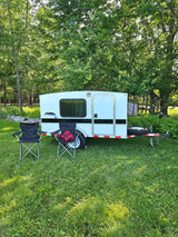 A white camper trailer, like an rv rental, with two folding chairs in a sunny, grassy spot under trees. Ready for your getaway!