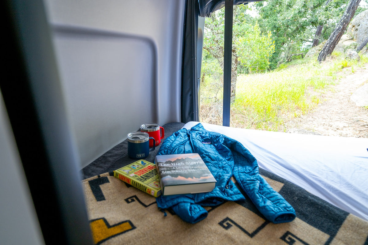 Enjoying an rv rental: books, mugs & a jacket on the bed, with a scenic window view of a green, wooded trail. — photo 8