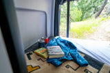 Enjoying an rv rental: books, mugs & a jacket on the bed, with a scenic window view of a green, wooded trail.