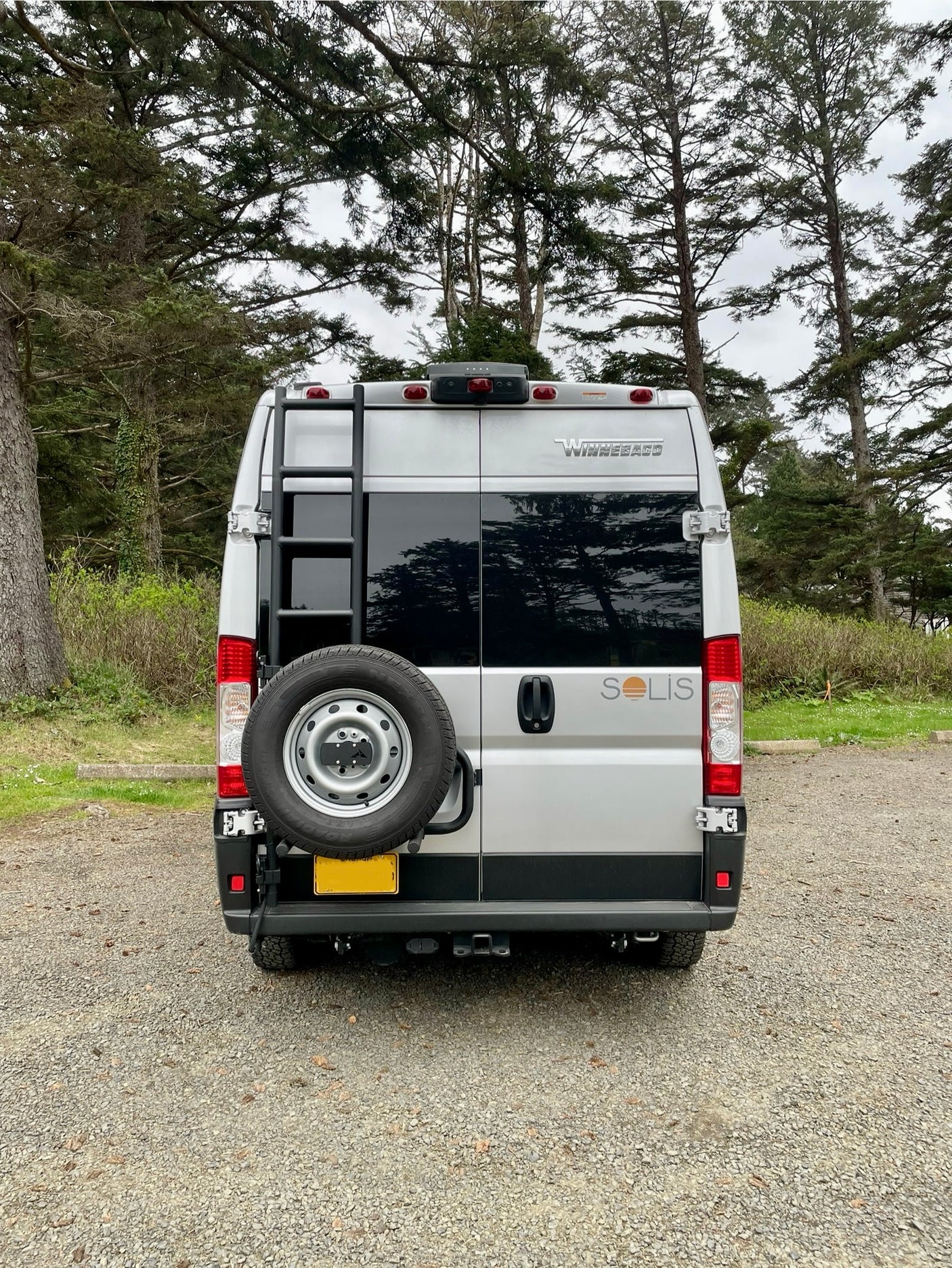 Silver Winnebago Solis camper van rv rental with spare tire and ladder, parked on gravel amidst tall trees.