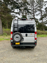 Silver Winnebago Solis camper van rv rental with spare tire and ladder, parked on gravel amidst tall trees.