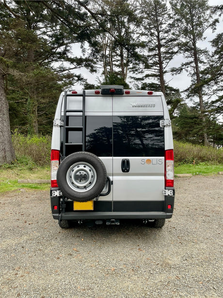 Silver Winnebago Solis camper van rv rental with spare tire and ladder, parked on gravel amidst tall trees.