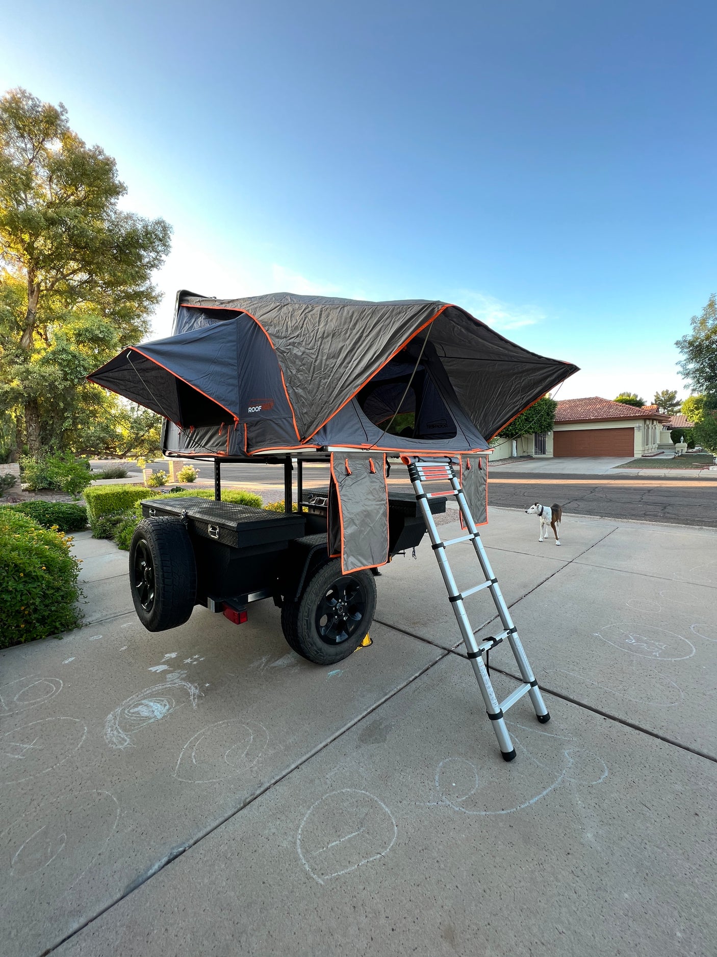 Grey rooftop tent on a trailer, fully deployed with ladder, spare tire & storage box. Dog nearby. Unique rv rental.