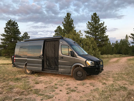 Adventure-ready grey RV rental van on a dirt path, surrounded by trees under a cloudy sky.
