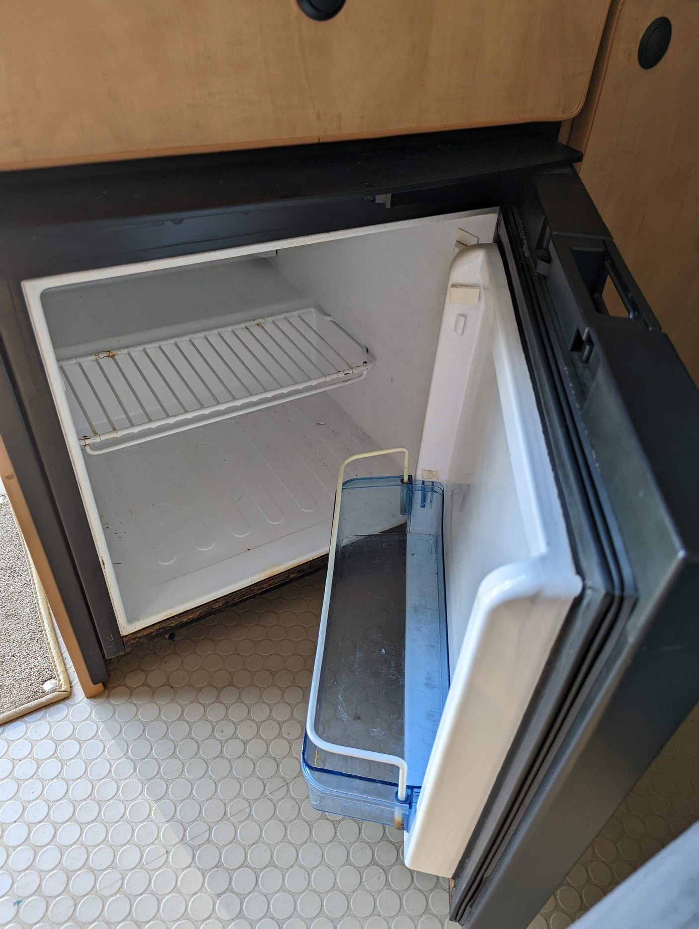 An empty, open RV fridge in an rv rental, showing its wire shelf and blue door bin. Needs cleaning. White, circular pattern floor.