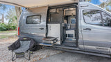 An open gray Winnebago rv rental shows an outdoor kitchen setup with griddle, camping chair, and extended awning, on gravel.