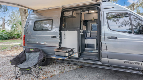 An open gray Winnebago rv rental shows an outdoor kitchen setup with griddle, camping chair, and extended awning, on gravel.