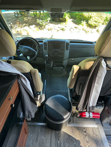RV rental camper van interior: From rear, view of front cockpit with tan seats, dashboard, and fire extinguisher. Green foliage outside.