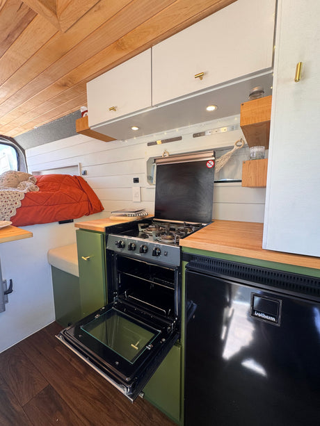 An RV rental's cozy kitchen area, showing an open oven, a refrigerator, green cabinets, and warm wood accents.