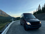 Experience nature with an rv rental! A dark camper van is parked on a mountain pass overlooking a scenic valley with a river and forests.