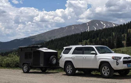 A white SUV with an off-road RV rental camper is perfect for mountain adventures. Scenic backdrop with forests & snowy peaks.