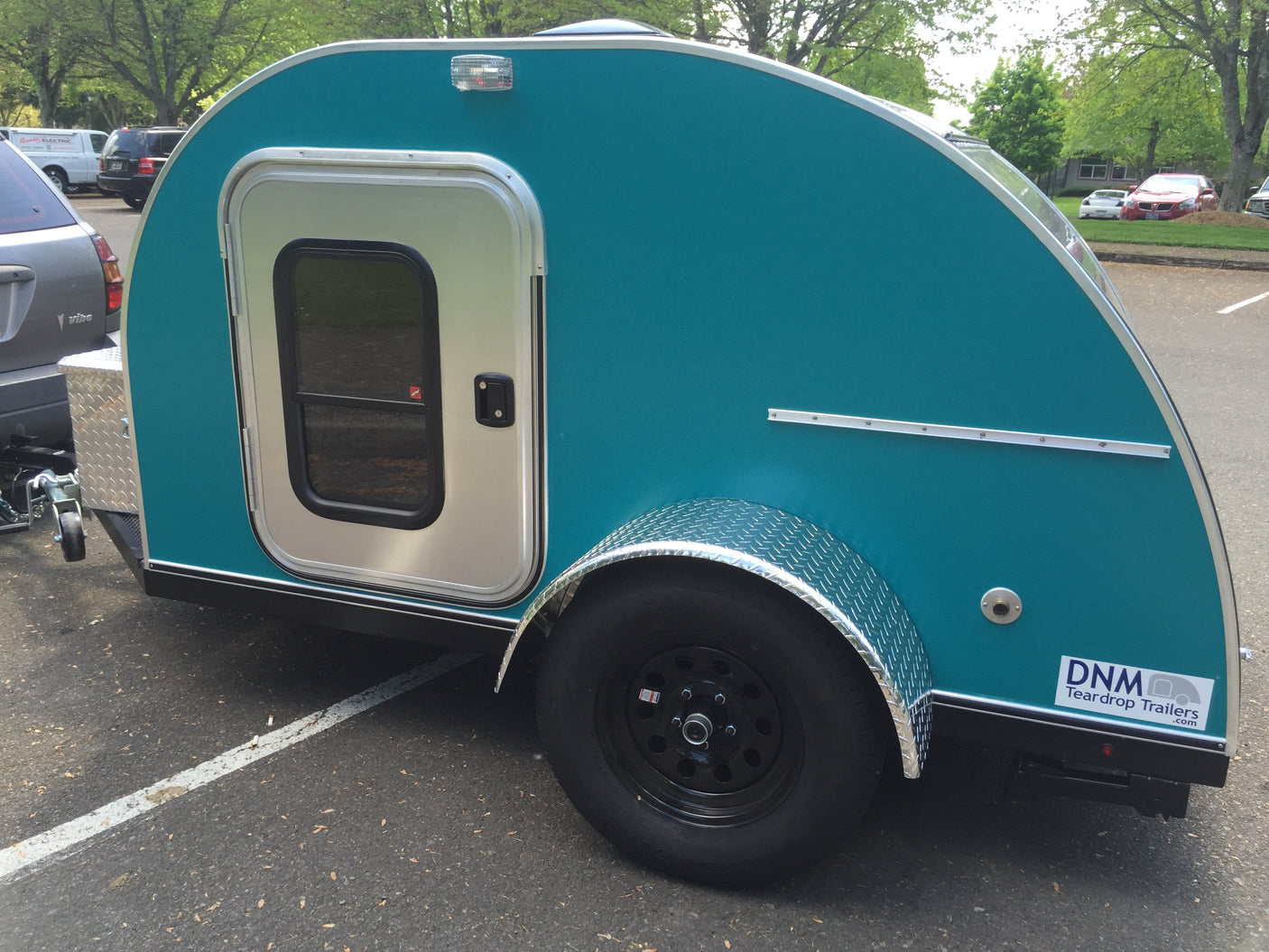 This vibrant teal teardrop RV rental, a compact camper, is parked on asphalt, featuring a side door and diamond plate fender.