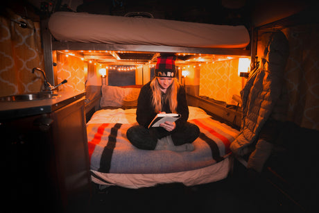 Cozy RV rental interior: A woman reads on a comfortable bed under warm string lights, featuring a bunk bed above and small sink area.