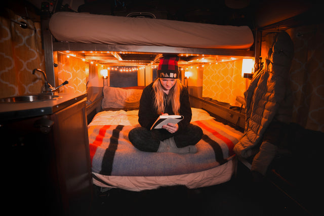 Cozy RV rental interior: A woman reads on a comfortable bed under warm string lights, featuring a bunk bed above and small sink area.