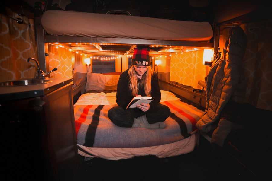 Cozy RV rental interior: A woman reads on a comfortable bed under warm string lights, featuring a bunk bed above and small sink area.