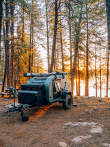 Rugged teardrop RV rental parked in a sun-drenched pine forest at golden hour, perfect for lakeside camping adventures.