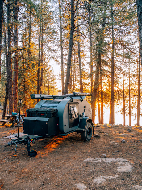 Rugged teardrop RV rental parked in a sun-drenched pine forest at golden hour, perfect for lakeside camping adventures.