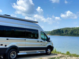 A light-colored RV rental van is parked beside a calm blue lake, surrounded by green trees under a clear sky.
