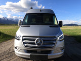 Silver Mercedes-Benz Sprinter camper van RV rental on a gravel road, backed by majestic snow-capped mountains and green fields.