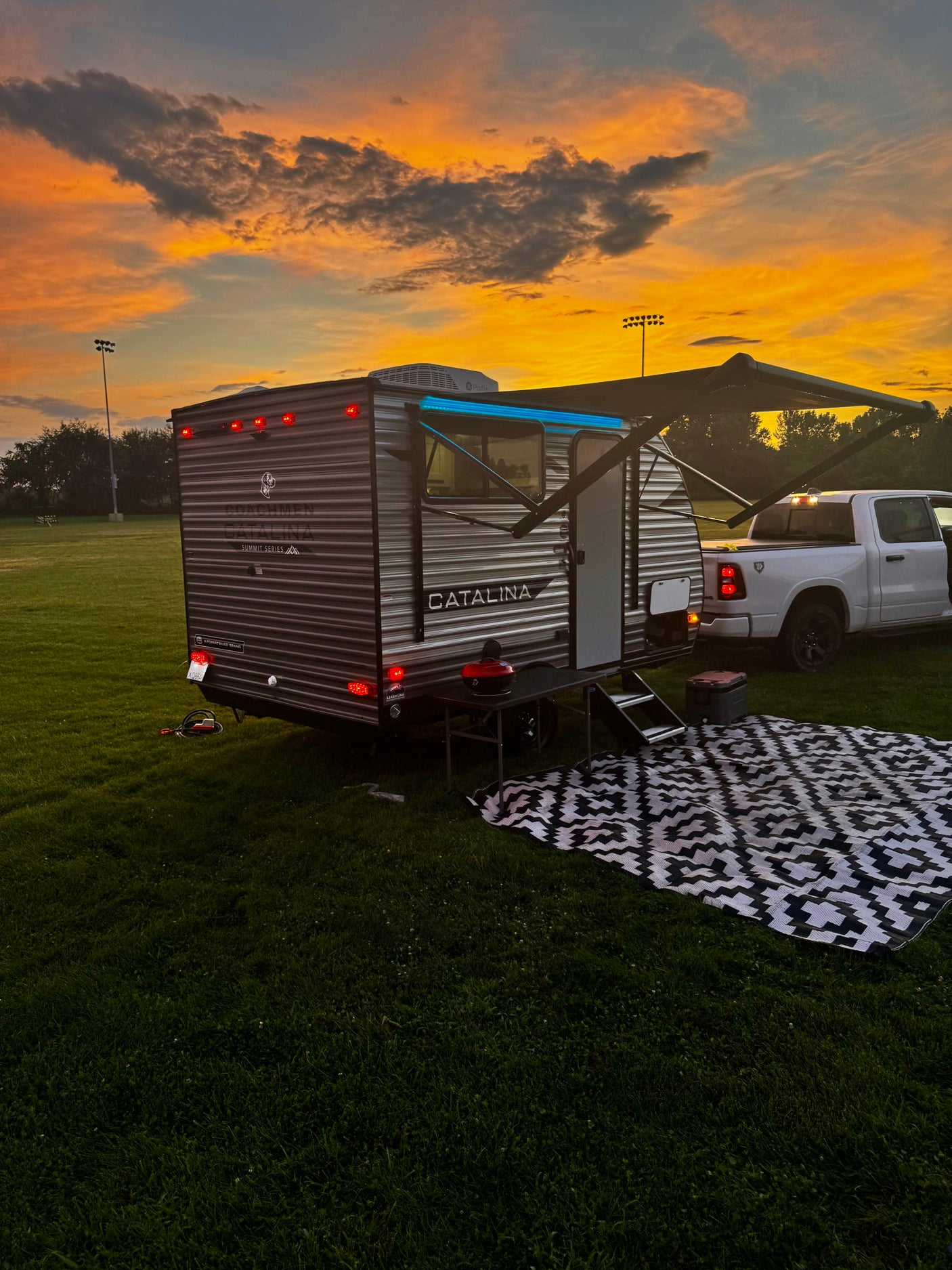 This RV rental offers a perfect outdoor setup: a travel trailer & truck on grass with a mat, under a vibrant sunset.