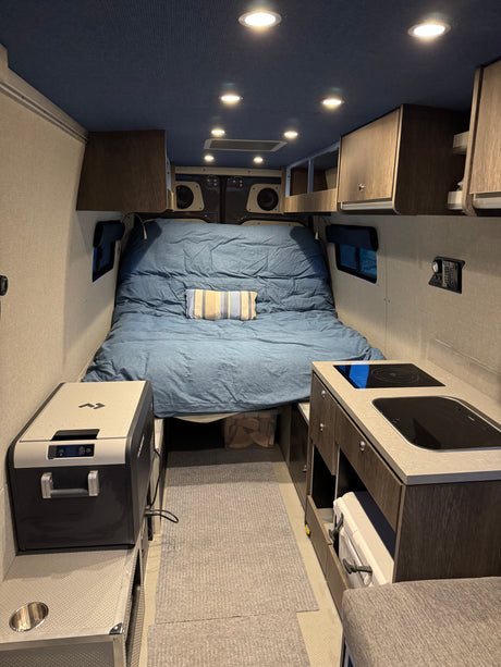 This rv rental interior features a comfortable bed, compact kitchen with cooktop, and a portable fridge, all within a well-lit space.