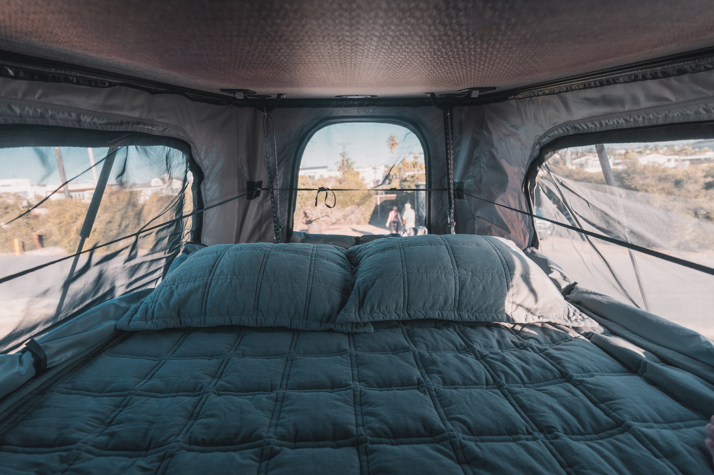 Cozy rv rental interior: A quilted bed with pillows inside a rooftop tent, offering scenic outdoor views through screened windows.