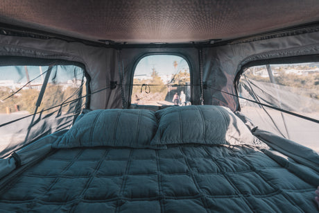 Cozy rv rental interior: A quilted bed with pillows inside a rooftop tent, offering scenic outdoor views through screened windows.