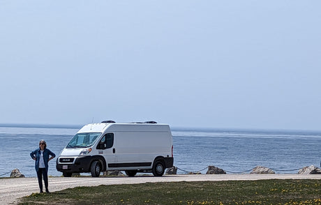 A white RAM ProMaster van, likely an RV rental, is parked near a calm blue body of water. A person stands on the left.