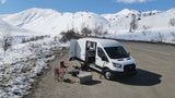 A white van, like an RV rental, is set up for winter camping in snowy mountains with open doors, chairs, and cooler.