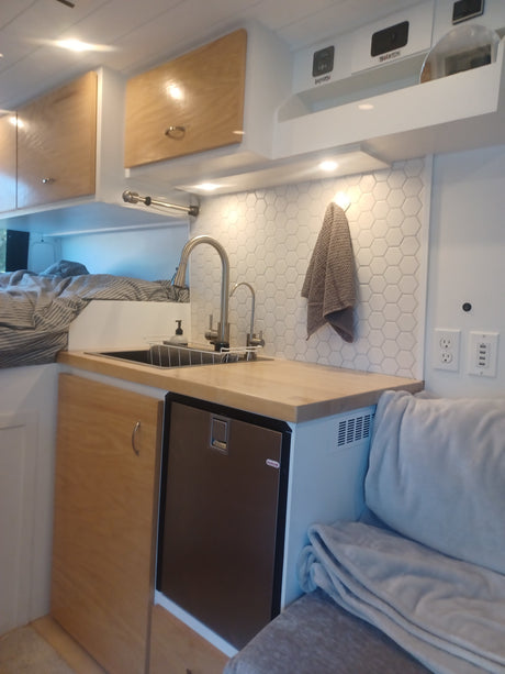 RV rental interior featuring a compact kitchen with sink, fridge, wood cabinets, and a bed. Controls for battery/inverter visible.