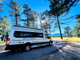 This silver rv rental camper van is parked on a road surrounded by sunny pine trees & distant mountains, ready for adventure!