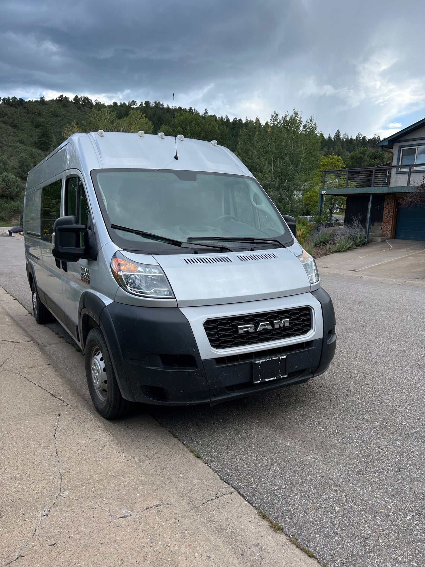 This silver Ram ProMaster camper van, available as an RV rental, is parked on a street with a wooded hillside.