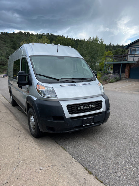 This silver Ram ProMaster camper van, available as an RV rental, is parked on a street with a wooded hillside.